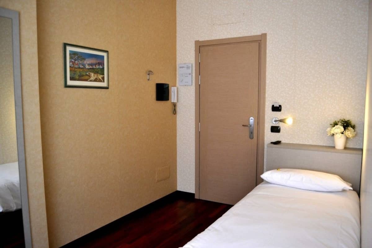 A hotel room with a wooden door, a small bedside table with a vase of flowers, and a framed picture on the wall.