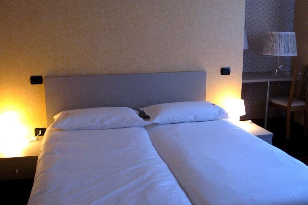 A double bed with white linens in a hotel room. There are two bedside tables with lamps, a chair, and a patterned wall.
