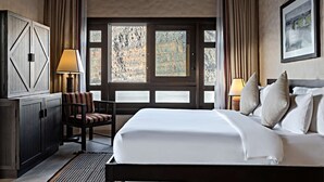 Habitación de hotel con una cama grande, un escritorio con lámpara, una silla y vistas a un paisaje montañoso por la ventana.