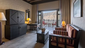 Habitación de hotel con un armario grande de madera oscura, un sofá a rayas, una mesita con una lámpara y vistas a un paisaje montañoso a través de las puertas del balcón.