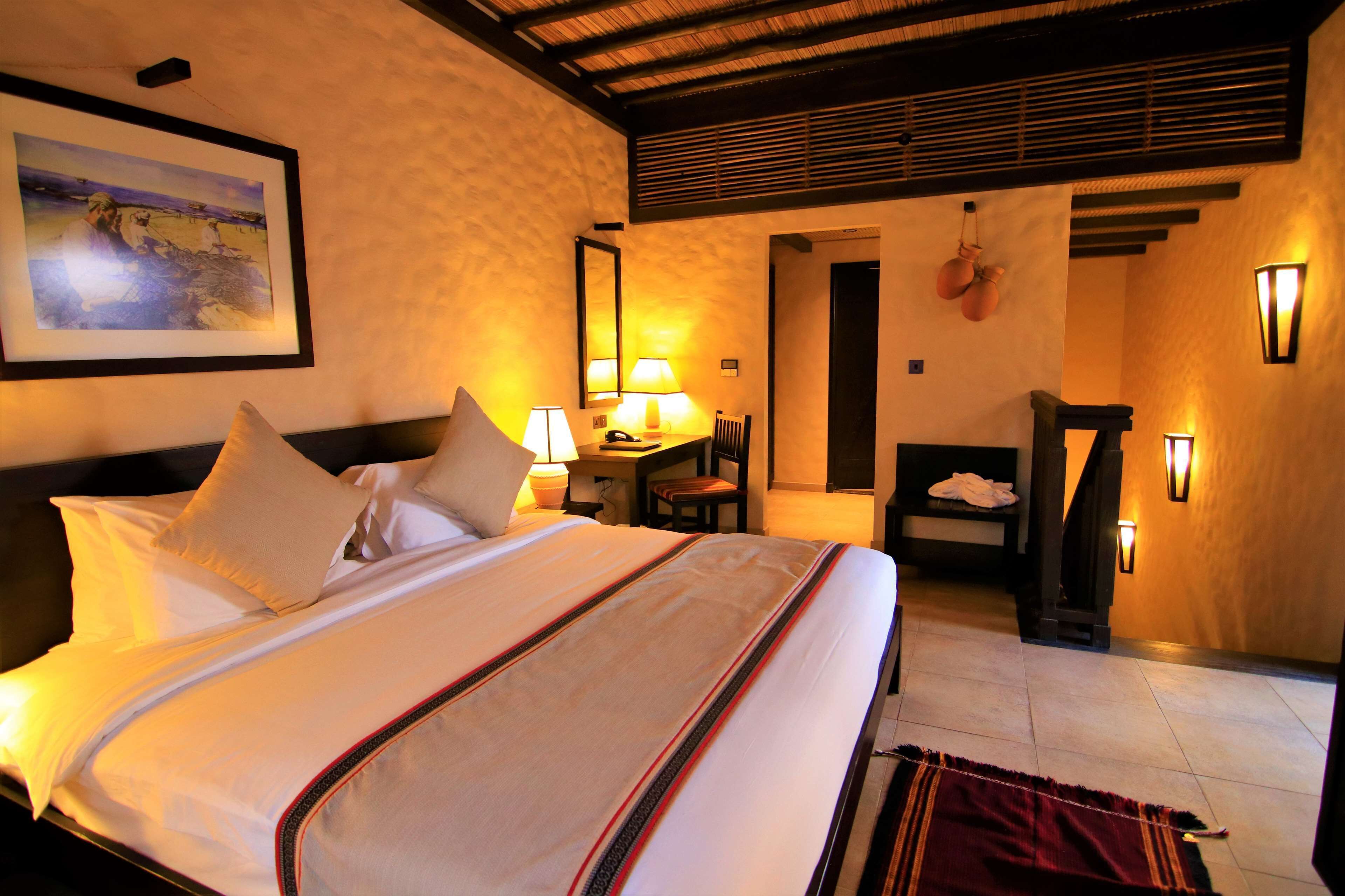 A hotel room with a bed, a desk, a chair, and a wall-mounted lamp.