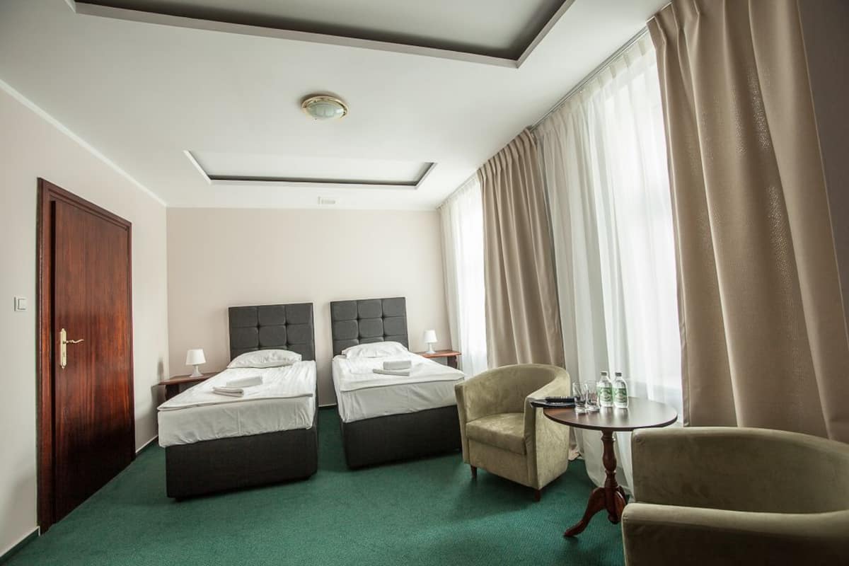 Ein Hotelzimmer mit zwei Betten, einem Sessel, einem kleinen Tisch mit Flaschen und einem Fenster mit Vorhängen.