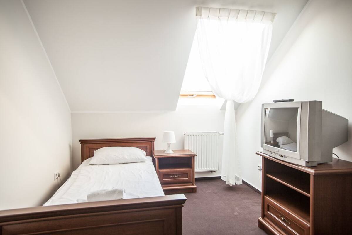 Ein kleines, helles Schlafzimmer mit einem Einzelbett, einem Nachttisch, einem Fernseher auf einem Holzständer und einem Fenster mit Vorhängen.