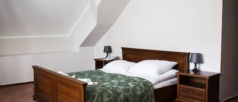 A bedroom with a wooden bed, green patterned bedspread, white pillows, two bedside tables with lamps, and a white wall.