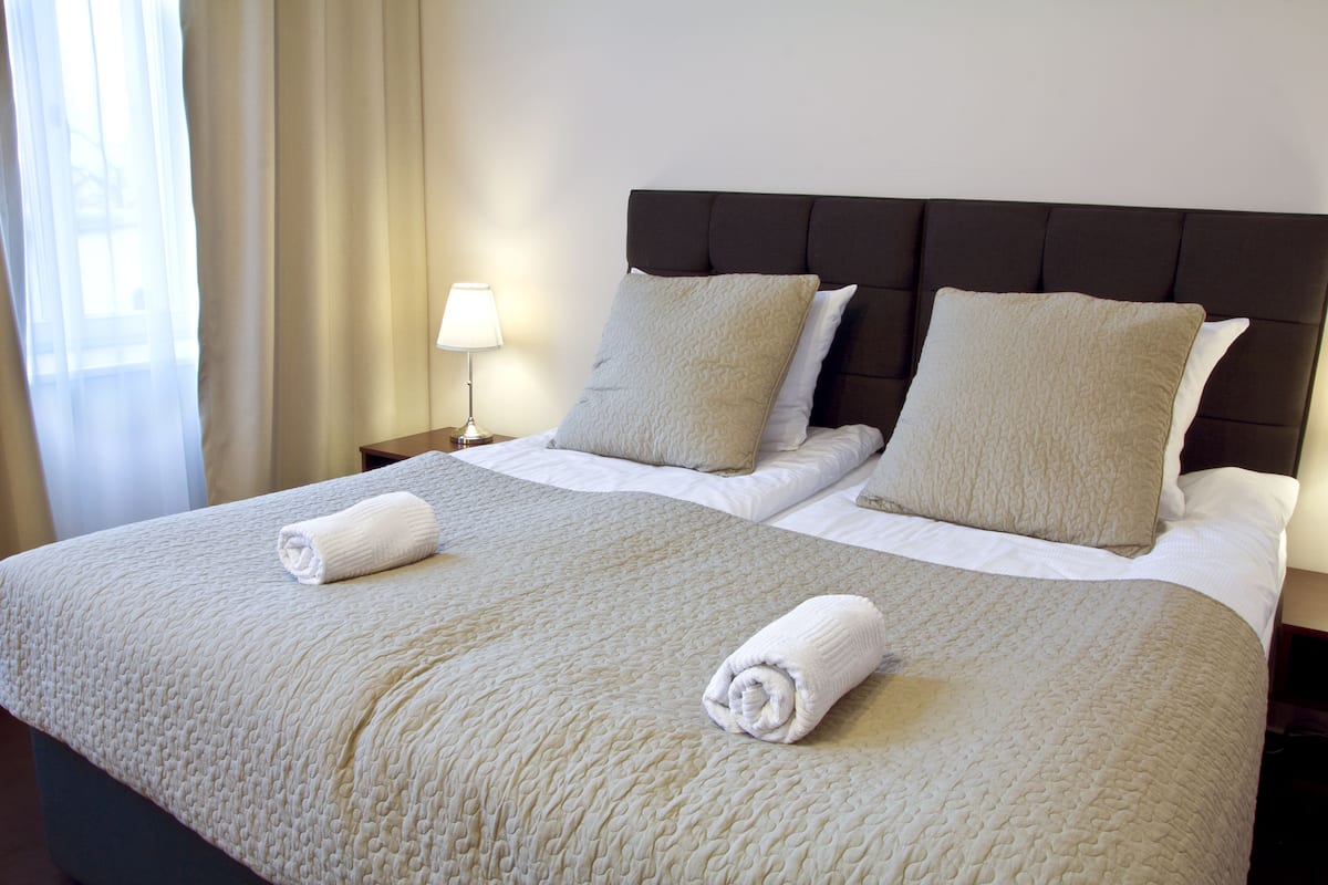 Ein ordentlich bezogenes Doppelbett mit einer beigen Tagesdecke, zwei Kissen und zwei aufgerollten Handtüchern an einem dunklen Kopfteil.