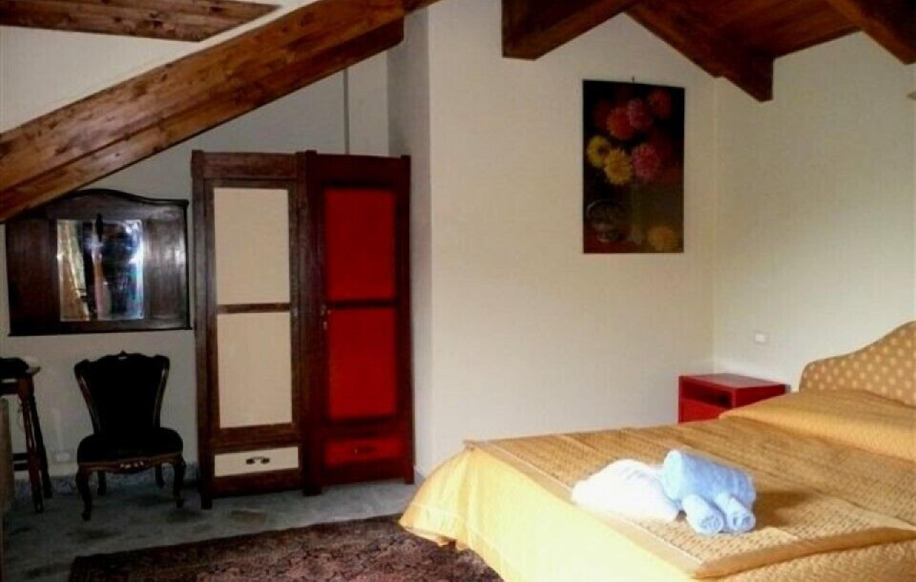 Un dormitorio con techo inclinado, una cama con colcha amarilla, una mesita de noche roja, una silla de madera y un cuadro en la pared.
