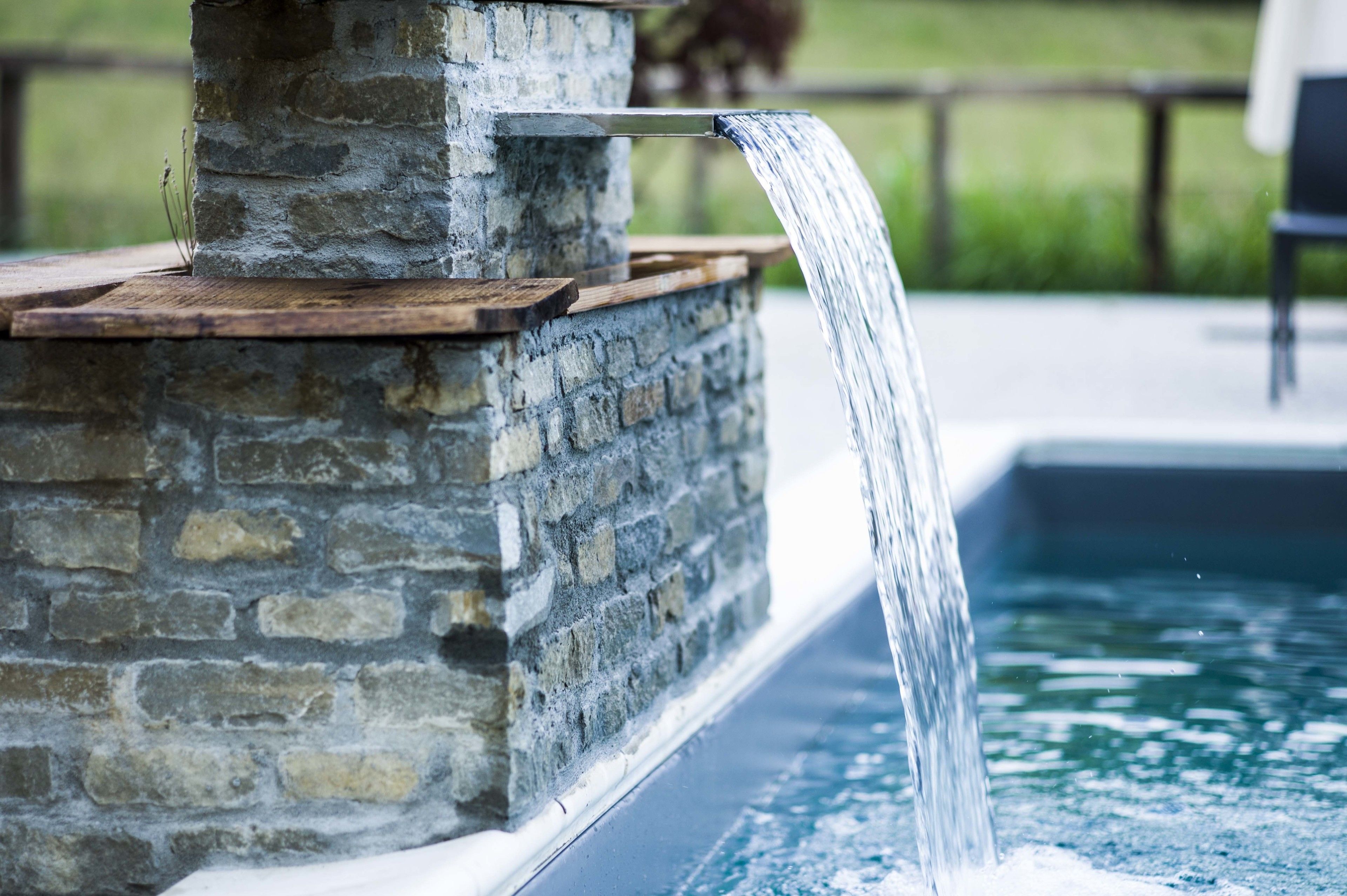Una fuente de piedra con un borde de madera y un caño de metal, que desemboca en una piscina.