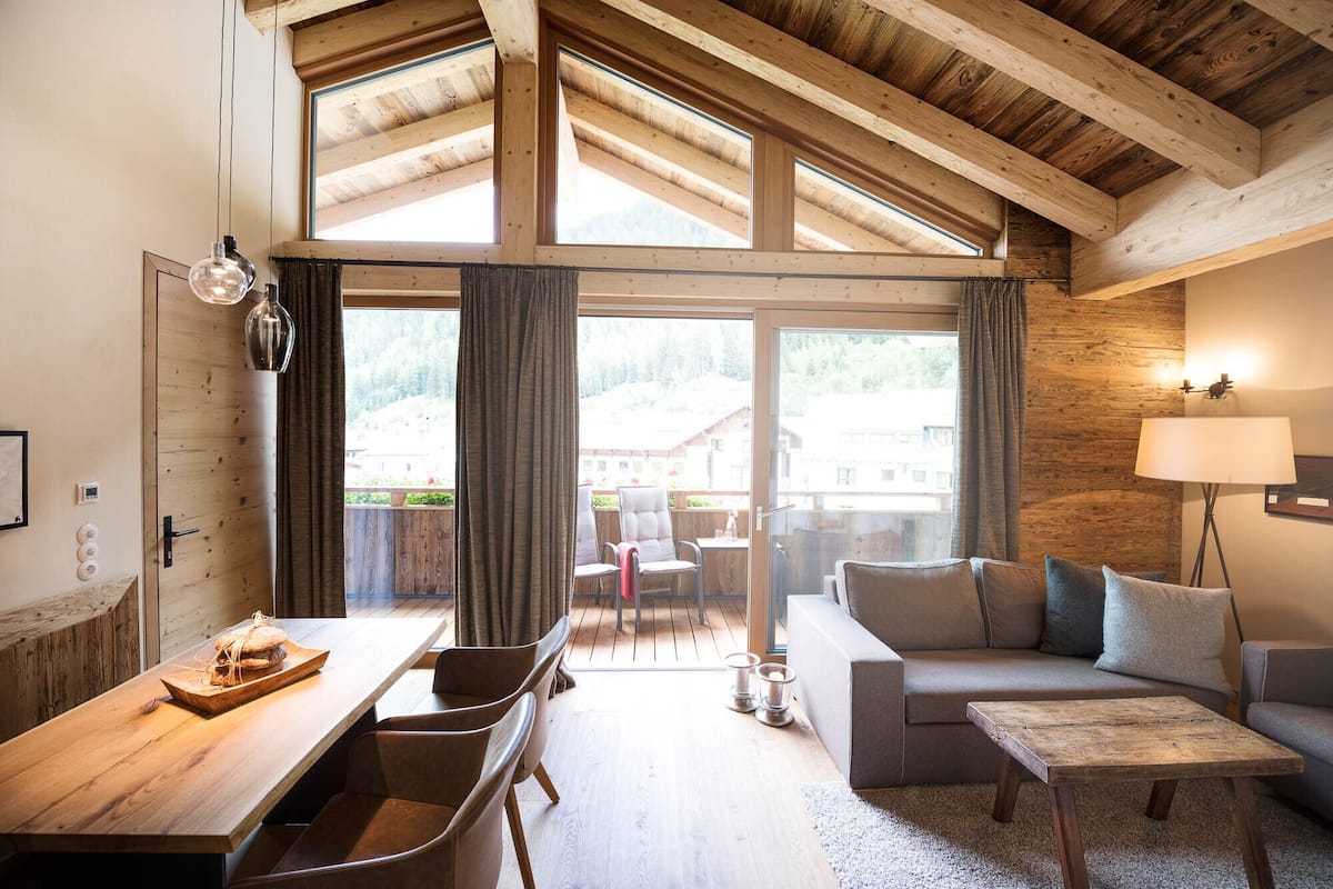 A spacious living area with a wooden ceiling, large windows, a sofa, a coffee table, and a dining table with chairs.