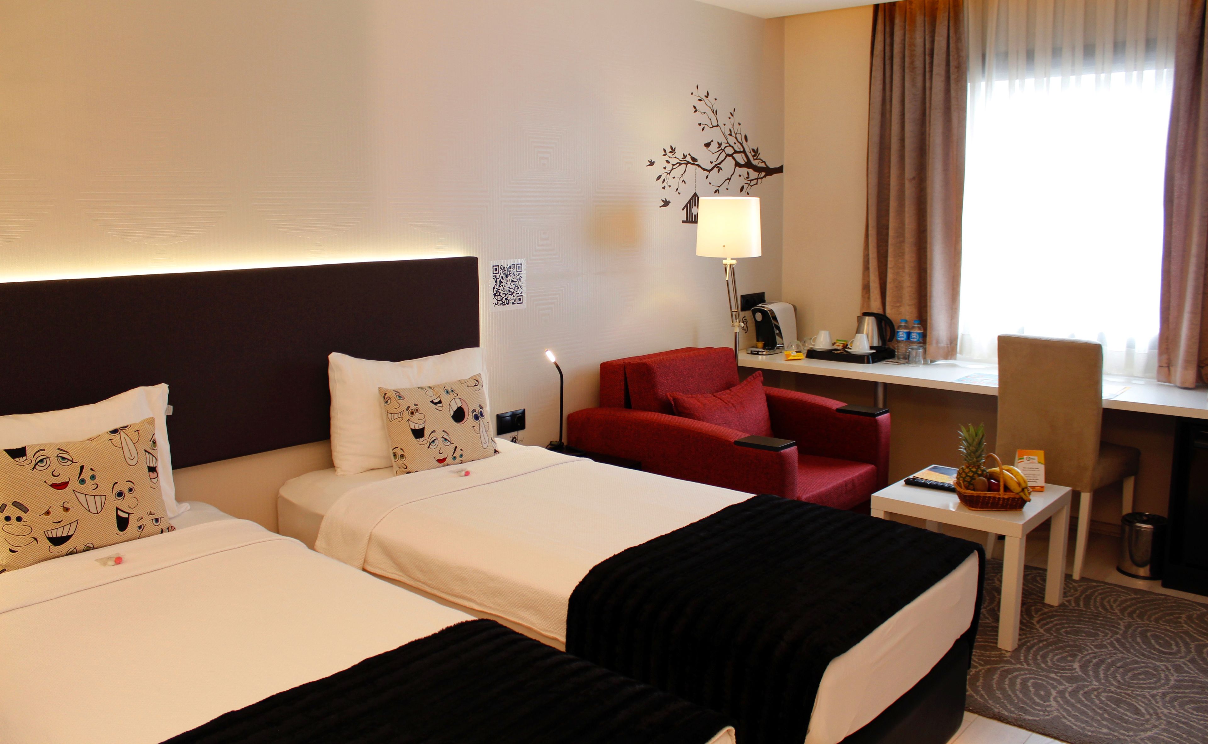 Habitación de hotel con dos camas, un escritorio, una silla, una lámpara y una pequeña mesa con una piña y plátanos.