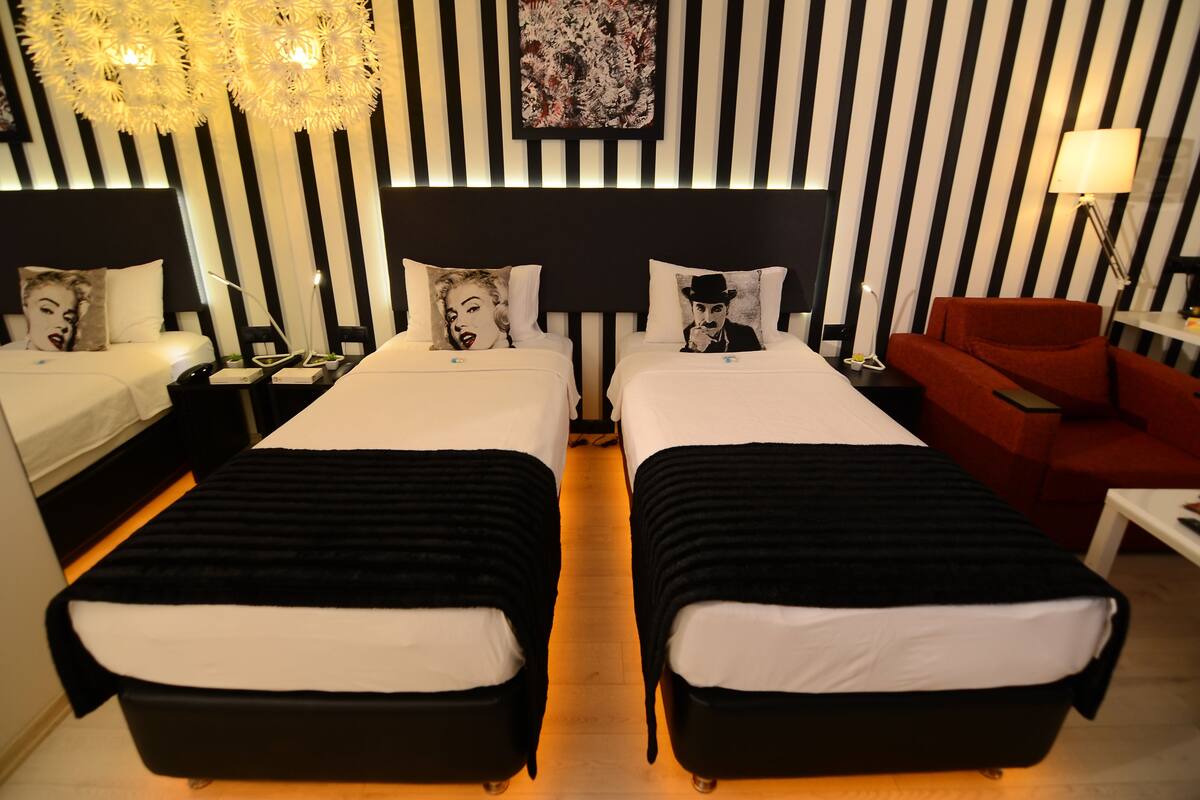 A hotel room with two beds, a red armchair, a nightstand with a lamp, and a wall with vertical stripes.