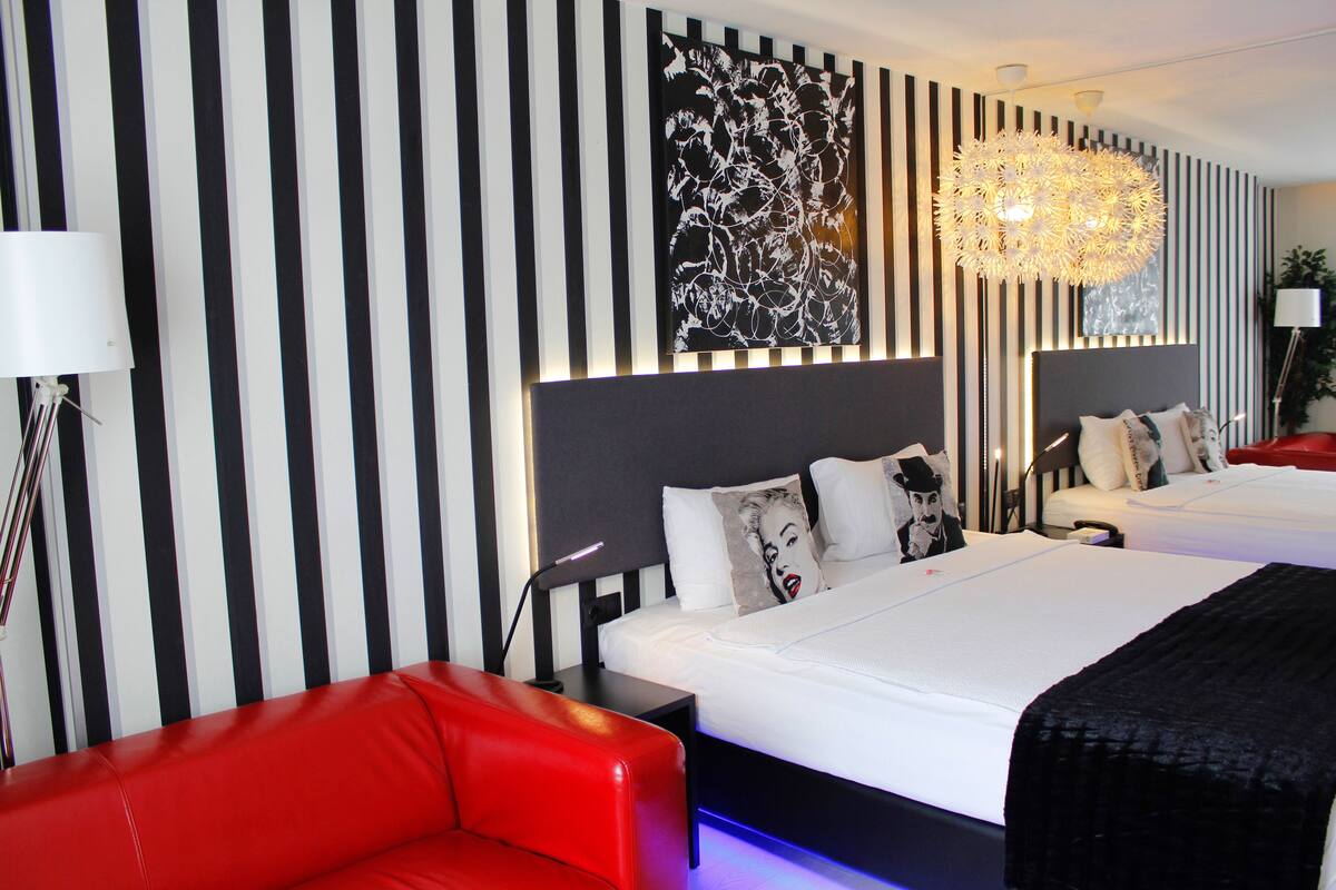 A hotel room with striped wallpaper, a large bed with white bedding, a red sofa, and a modern chandelier.