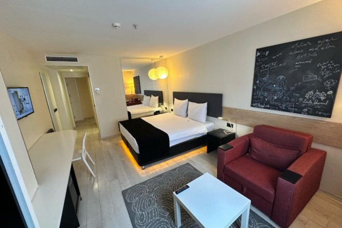 Una habitación de hotel con una cama, mesitas de noche, un sillón rojo, una pequeña mesa blanca y una pared con una pizarra.
