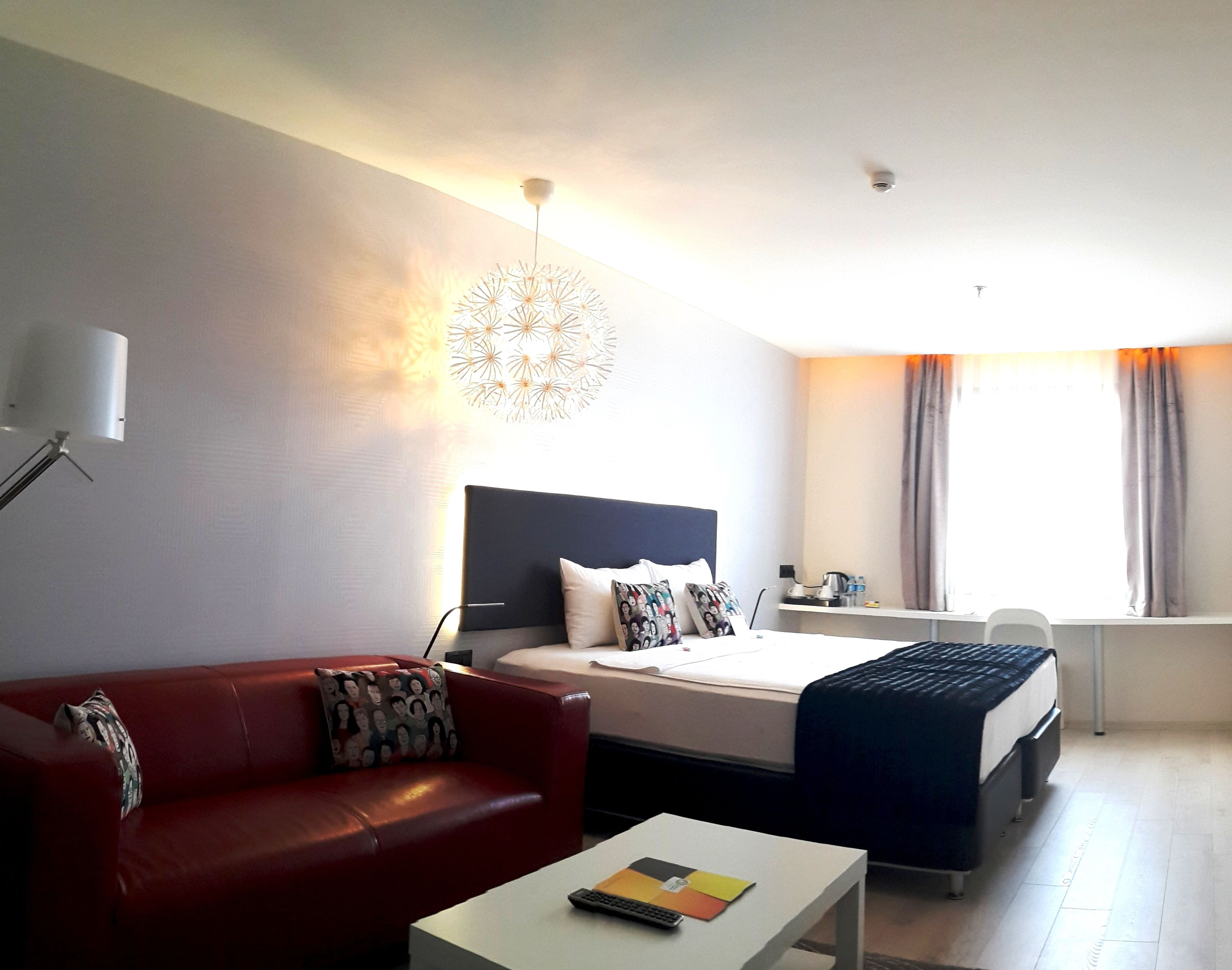 A hotel room with a bed, a red sofa, a white coffee table, and a modern chandelier.