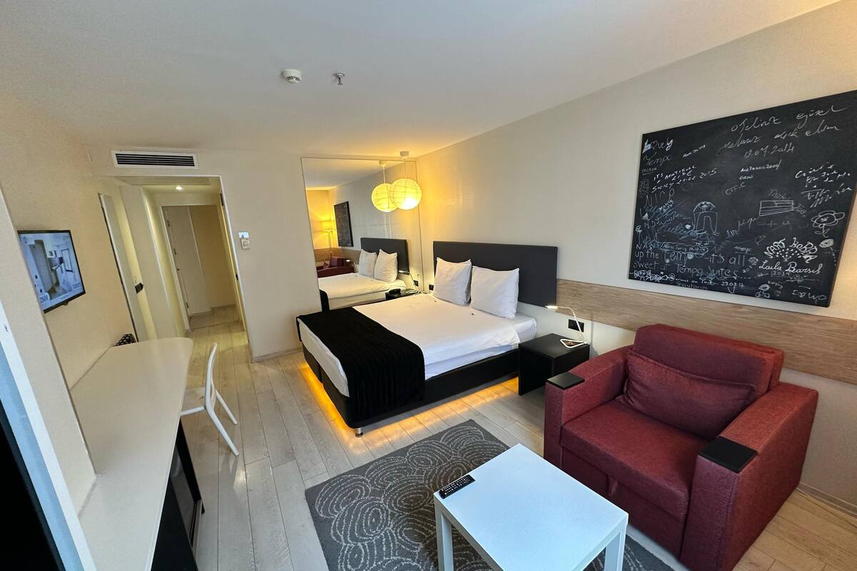 A hotel room with a bed, bedside table, chair, and a wall with a chalkboard.