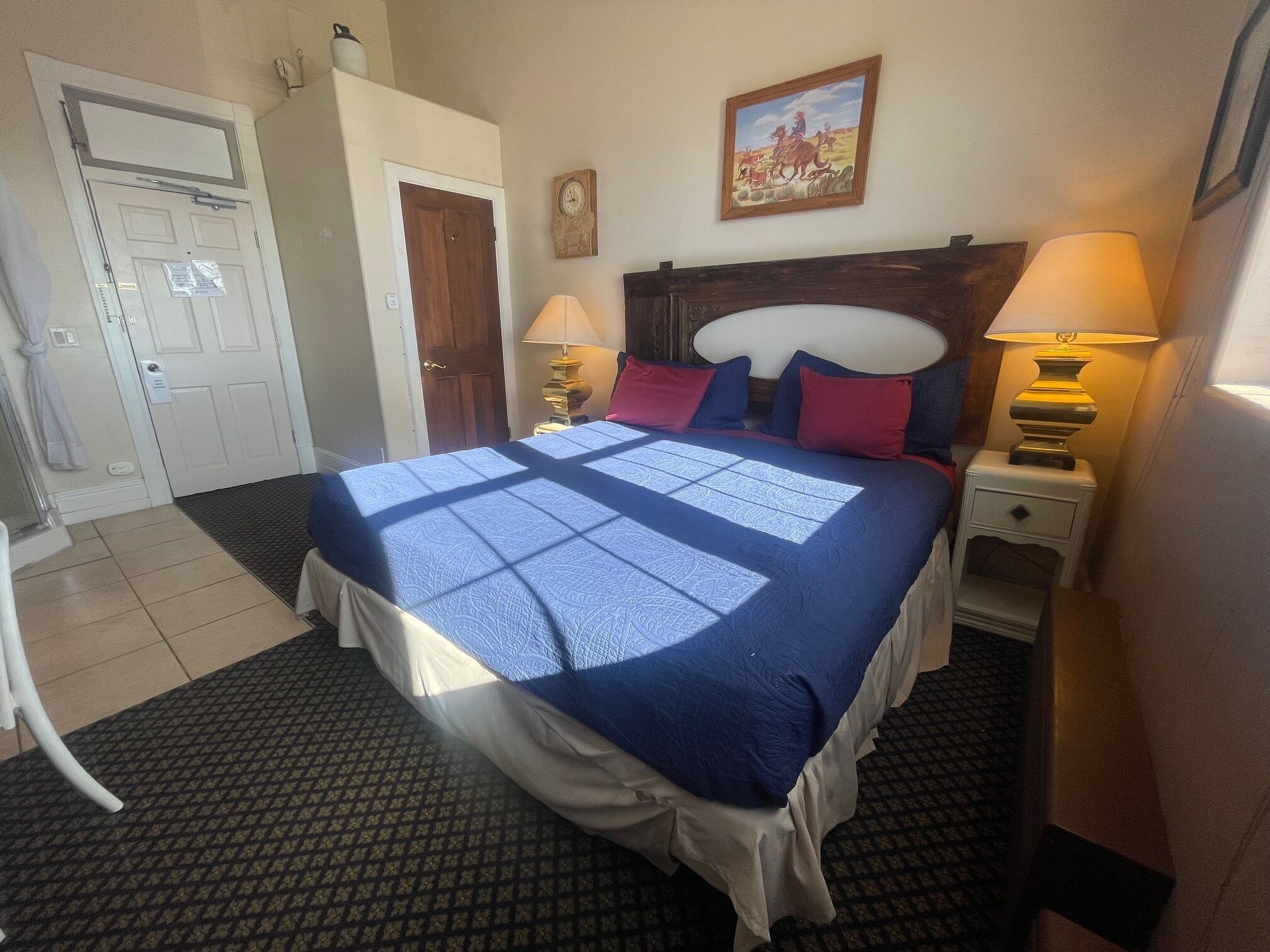 A hotel room with a large bed, two bedside lamps, a wooden headboard, a wall clock, and a framed picture above the bed.