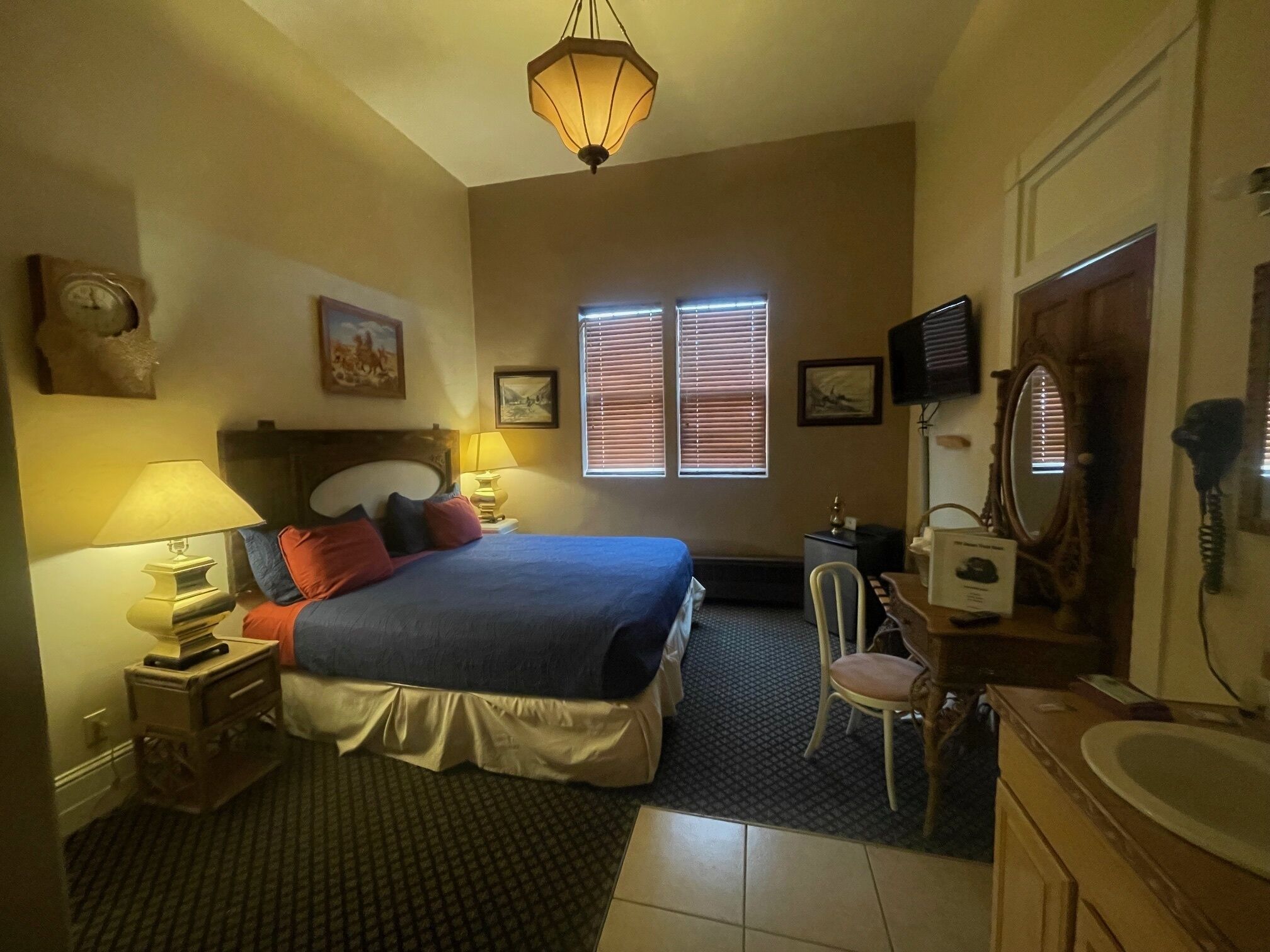A hotel room with a bed, two bedside lamps, a desk with a chair, a television, a mirror, and a window with blinds.