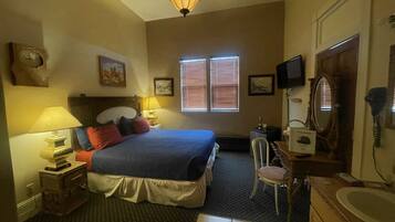 A hotel room with a bed, two bedside lamps, a desk with a chair, a television, a mirror, and a window with blinds.