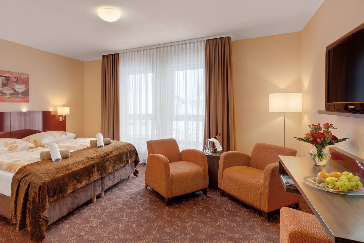 Habitación de hotel con una cama, dos sillones, un escritorio con frutero, un televisor y un cuadro en la pared.
