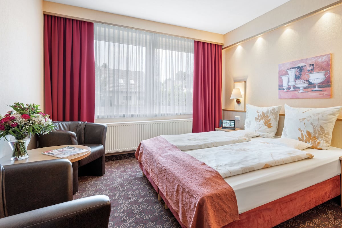 Habitación de hotel con dos camas, un escritorio con silla, ventana con cortinas rojas, un jarrón de flores y un cuadro en la pared.