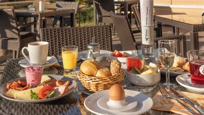 Un área de comedor al aire libre con muebles de mimbre, diversas opciones de desayuno y una botella de jugo de naranja Oerhof.