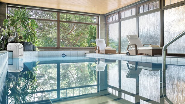 Una piscina con agua cristalina, rodeada de grandes ventanales que ofrecen vistas a árboles. Hay sillas de descanso y una pequeña mesa junto a la piscina.
