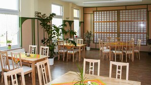 A dining area with wooden tables and chairs, potted plants, and large windows allowing natural light.