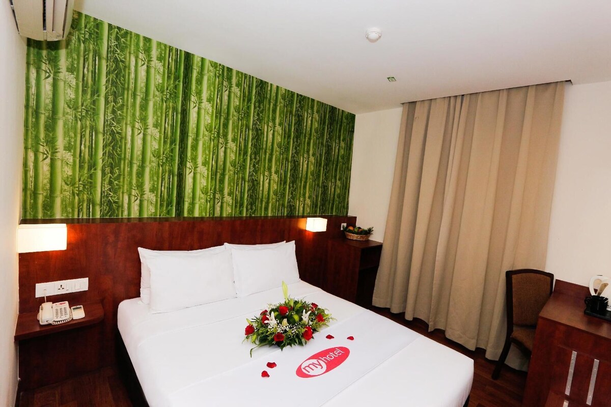 A hotel room with a bed, a nightstand, a desk, a chair, a wall with a bamboo wallpaper, and a window with curtains.