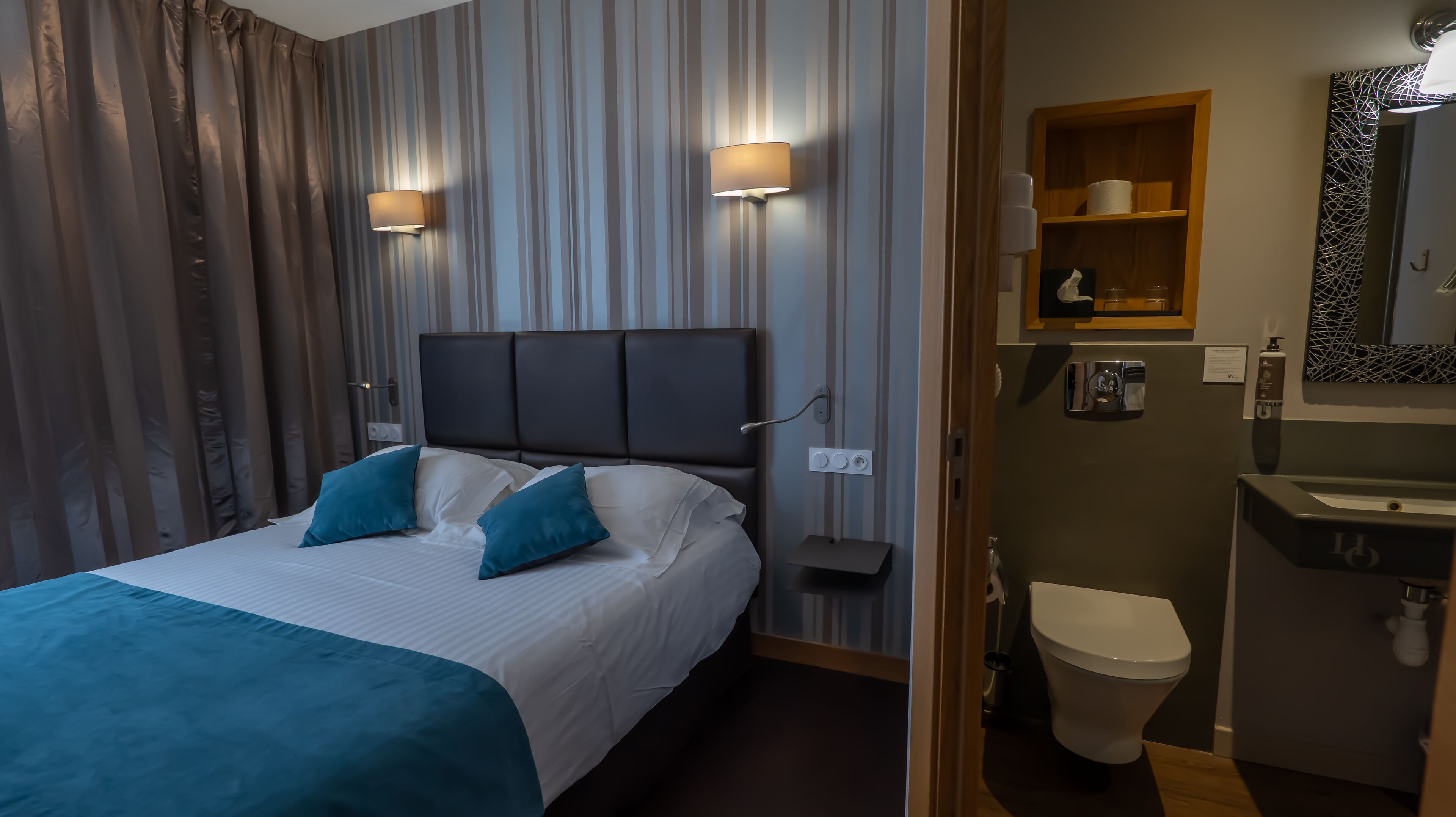 A hotel room with a bed, two wall-mounted lights, a bedside table, a mirror, a bathroom with a sink and toilet.