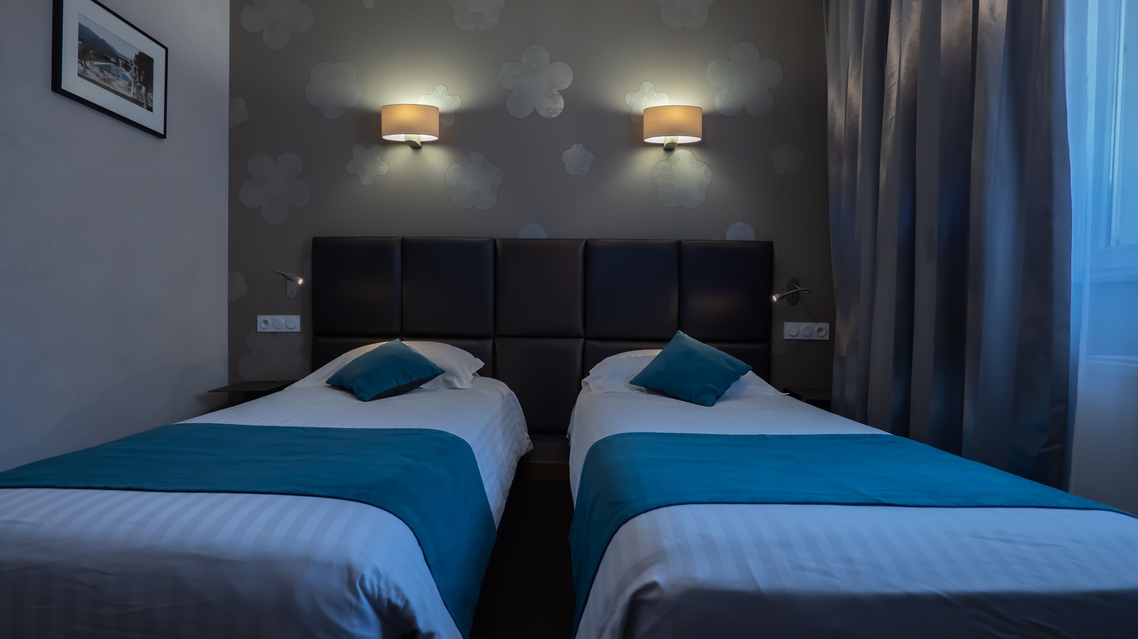 Habitación de hotel con dos camas, un cabecero oscuro y lámparas fijadas a la pared.
