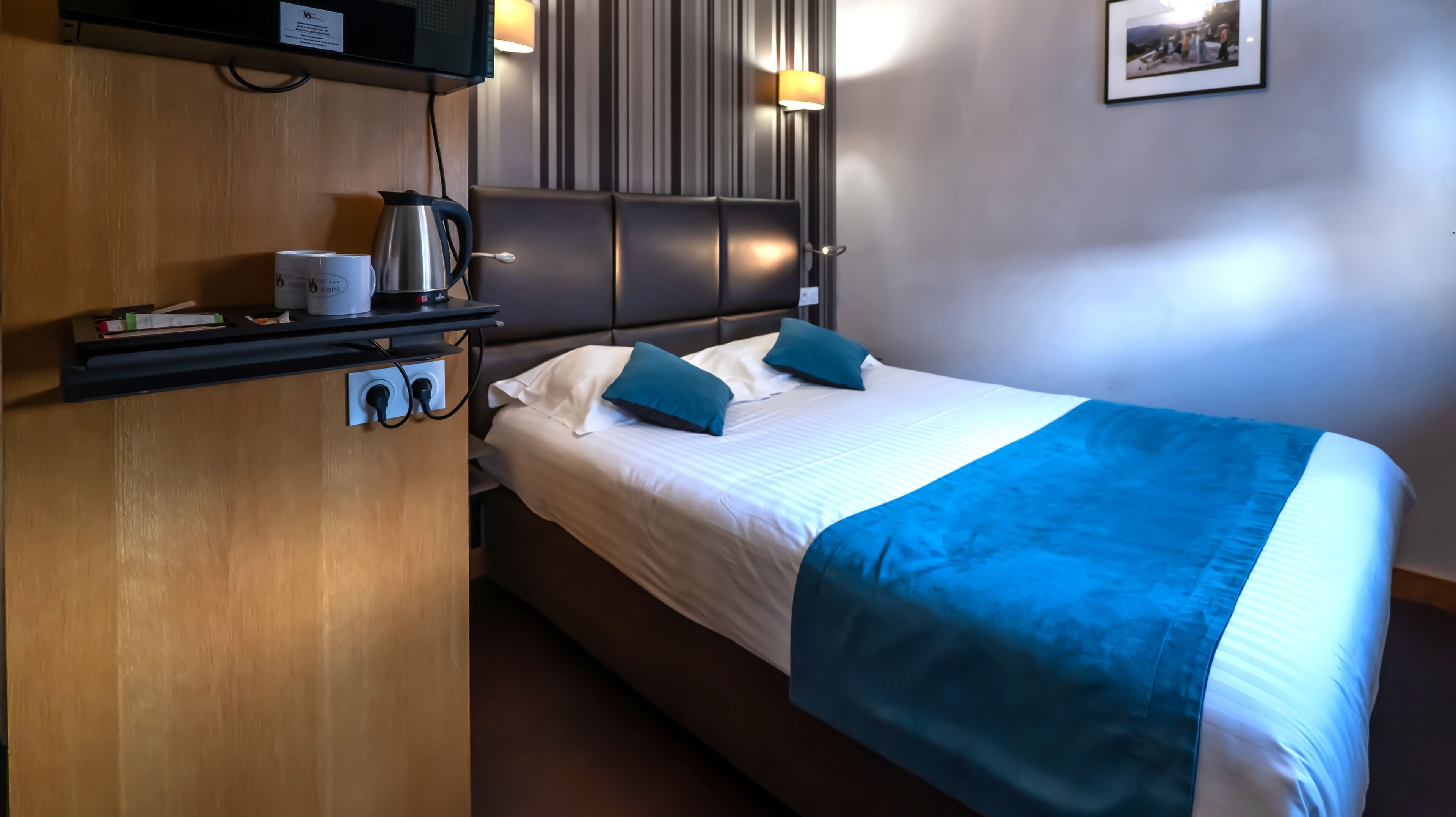 A hotel room with a bed, a bedside table with a kettle, a lamp, and a picture on the wall.