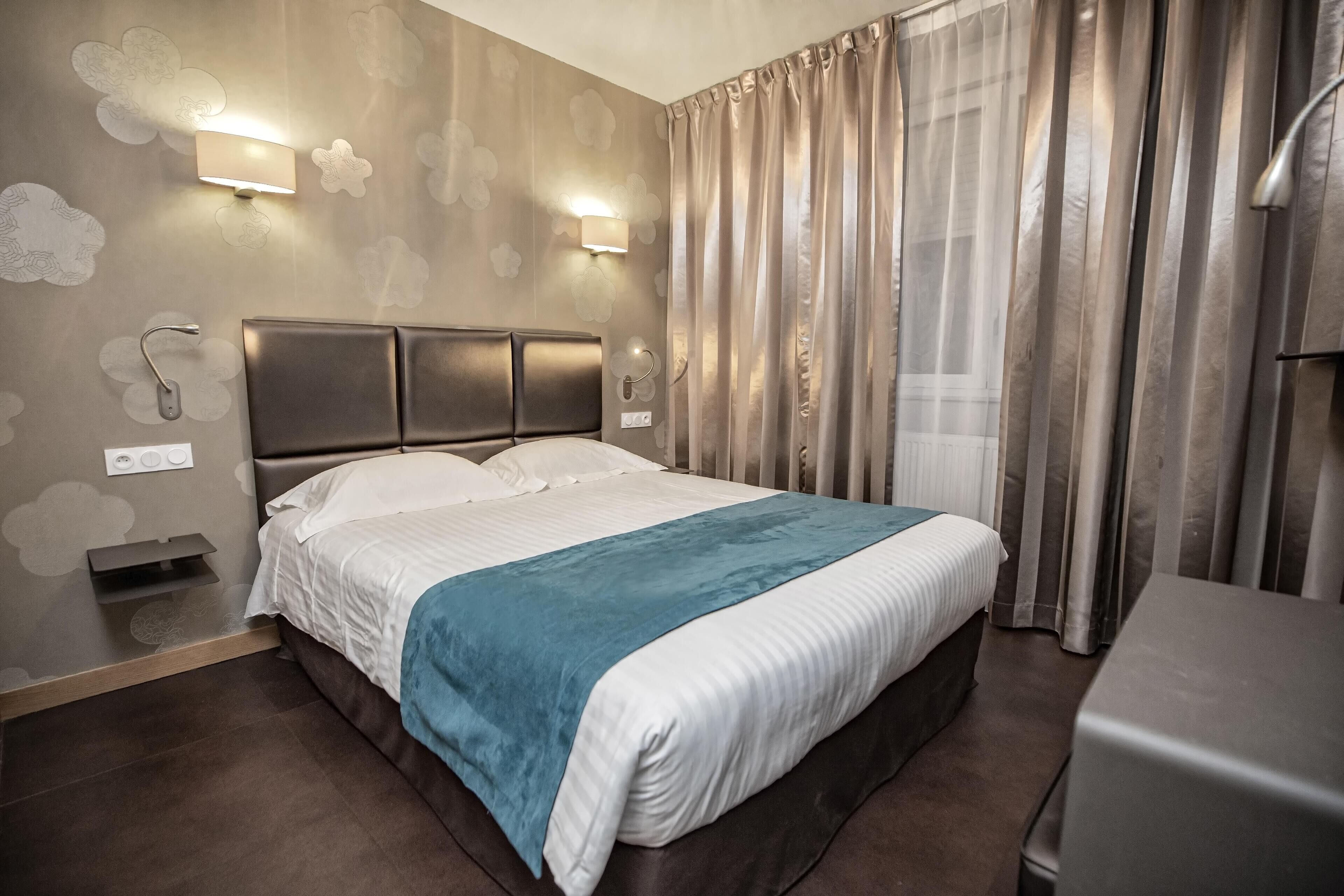 A hotel room with a large bed, a bedside table, a wall-mounted light, and a window with curtains.