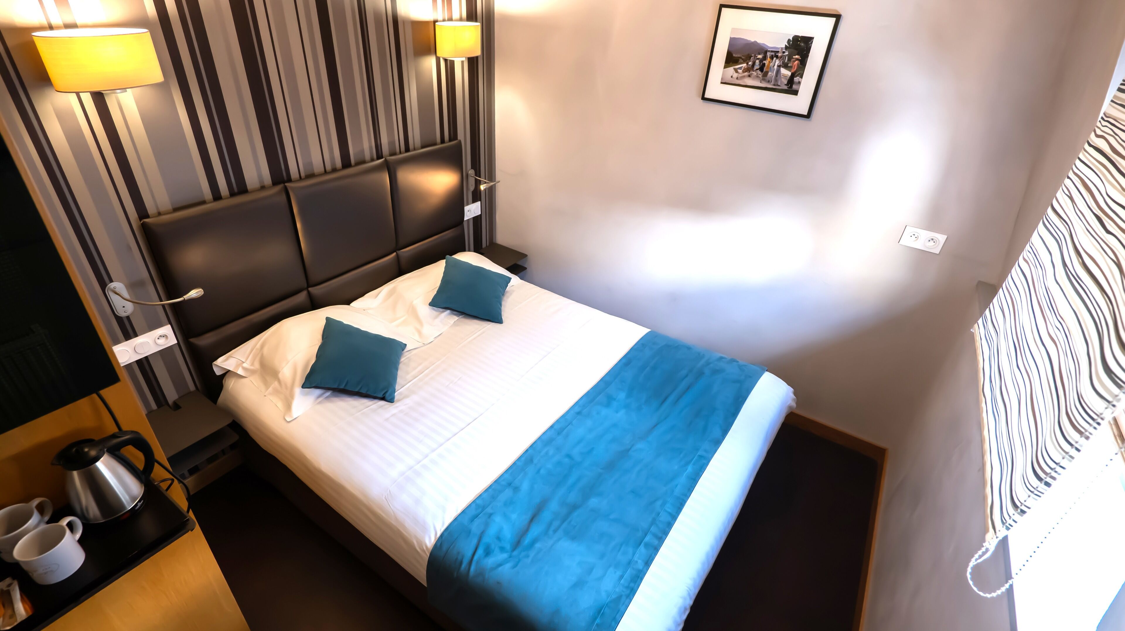 Habitación de hotel con cama, dos almohadas, mesita de noche con tetera y tazas, lámpara de pared e imagen enmarcada en la pared.