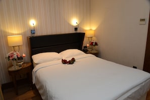 Una cama bien hecha con sábanas blancas, dos lámparas de noche y un florero con flores sobre una mesita de noche.