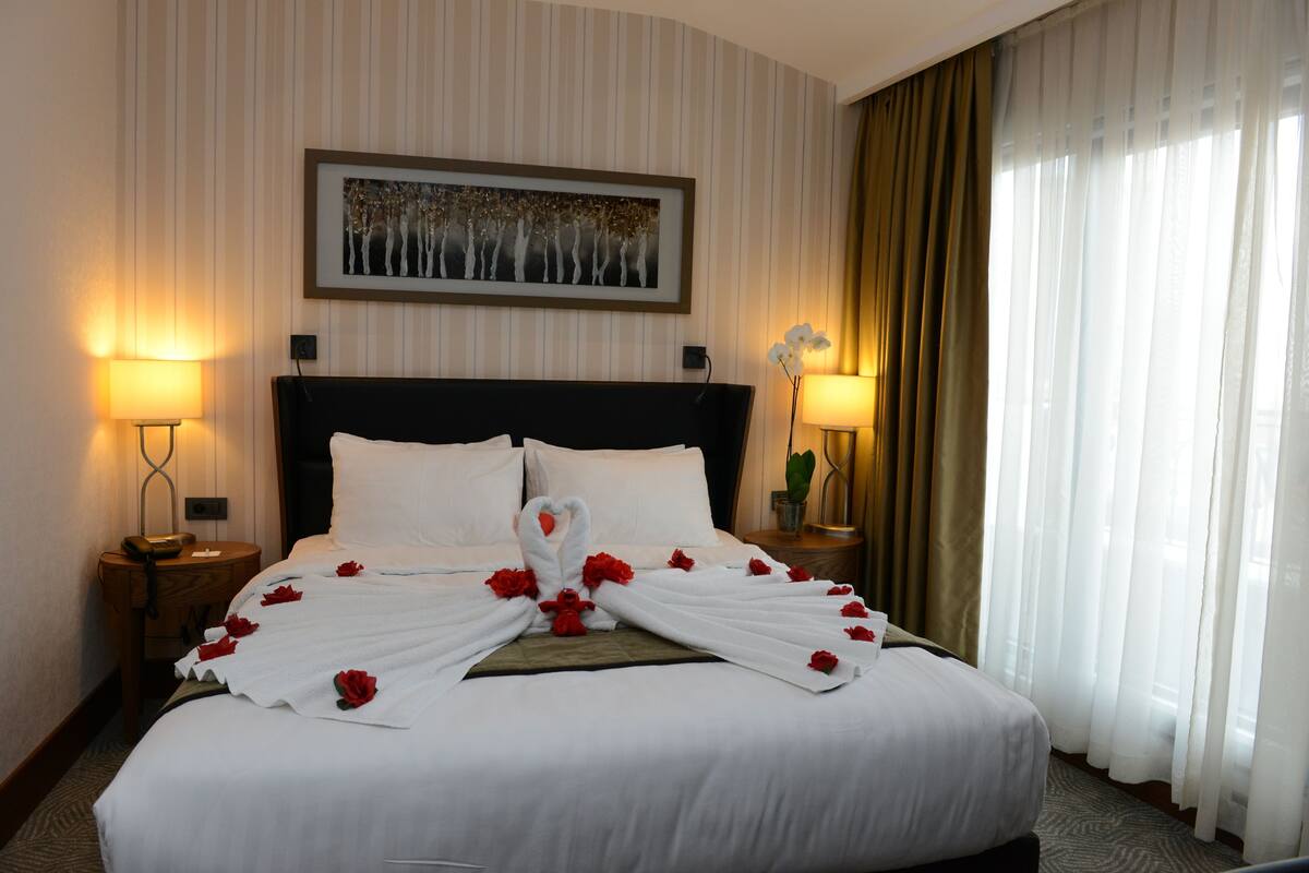 Una cama bien tendida con sábanas blancas y flores rojas, dos lámparas de noche, un cuadro en la pared y una ventana con cortinas.