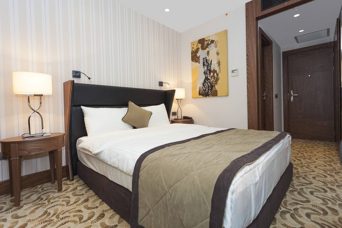 Habitación de hotel con una cama grande, dos mesitas de noche con lámparas, una puerta de madera y una obra de arte en la pared.