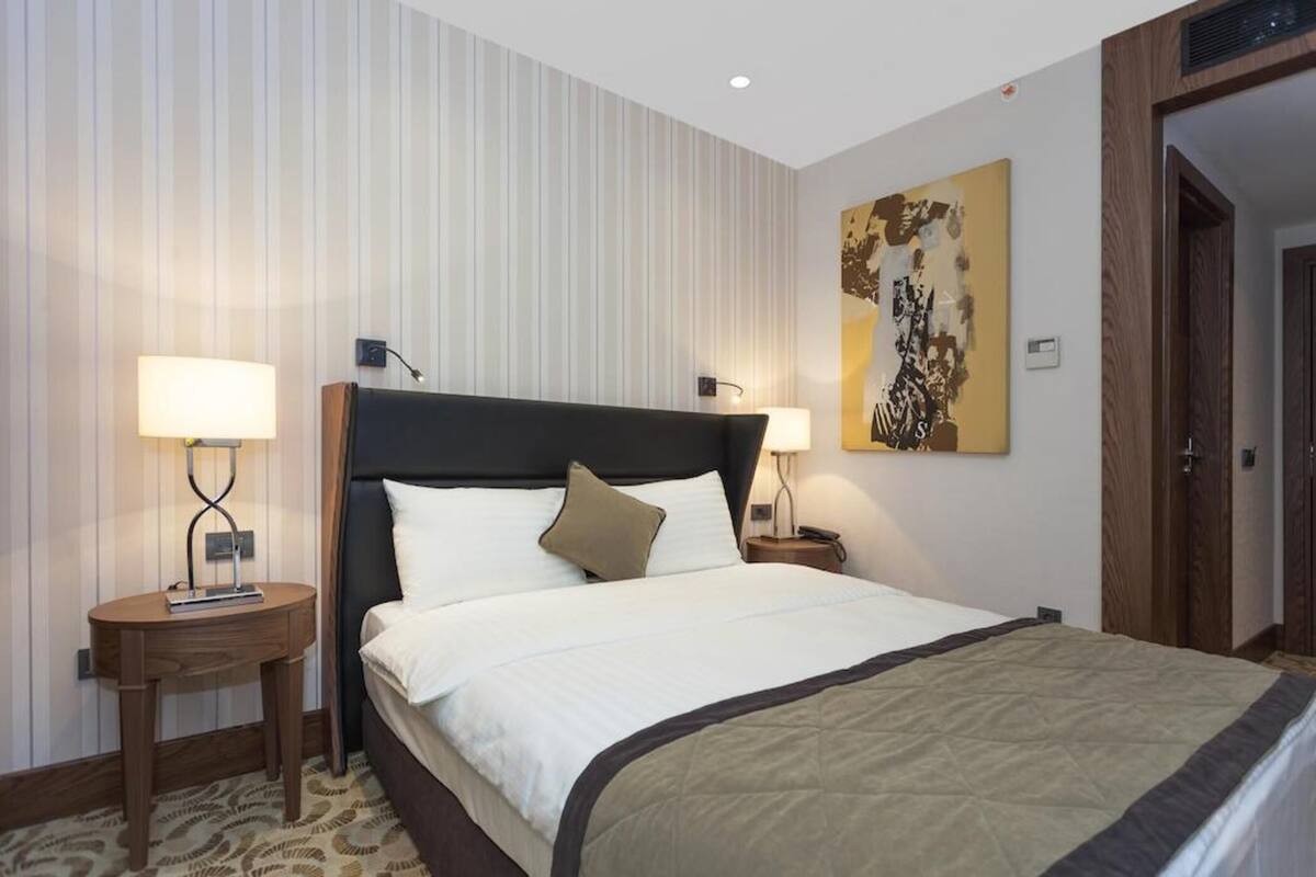 Habitación de hotel con una cama grande, dos mesitas de noche con lámparas, una obra de arte en la pared y una puerta de madera.