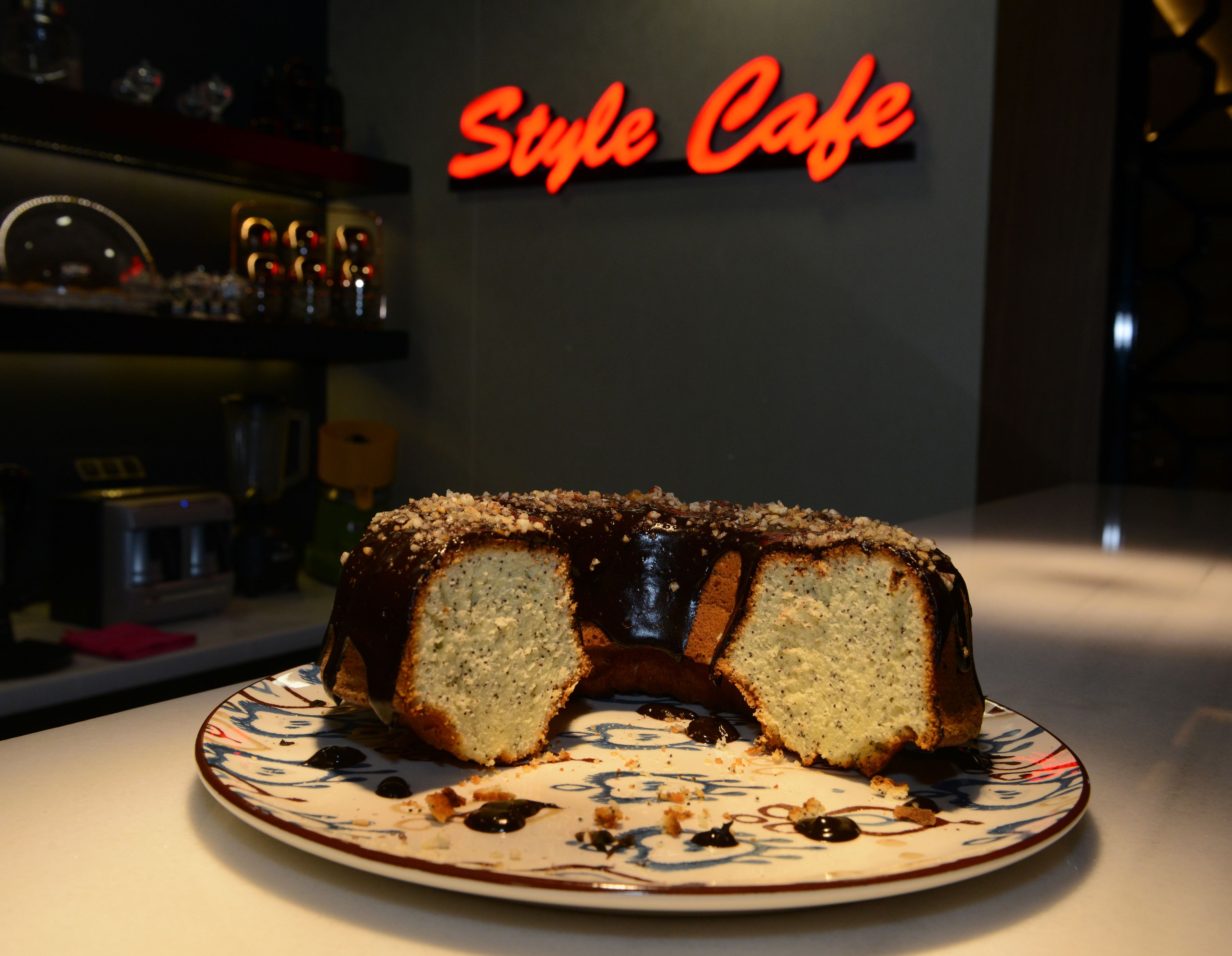 A slice of cake on a plate with a decorative pattern, placed on a table in a cafe with a neon sign reading "Style Cafe" in the background.