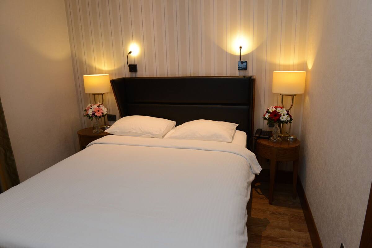 Una cama bien hecha con sábanas blancas, dos mesitas de noche con lámparas y arreglos florales.
