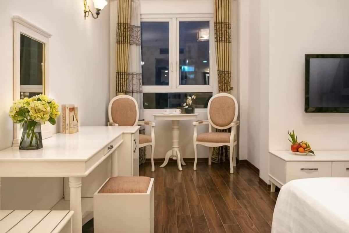 Et hotelværelse med en seng, et skrivebord med to stole, et lille bord, et fjernsyn og et vindue med gardiner.