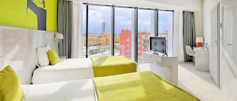 Habitación de hotel con dos camas, un escritorio con una computadora, un televisor y vistas a la ciudad.