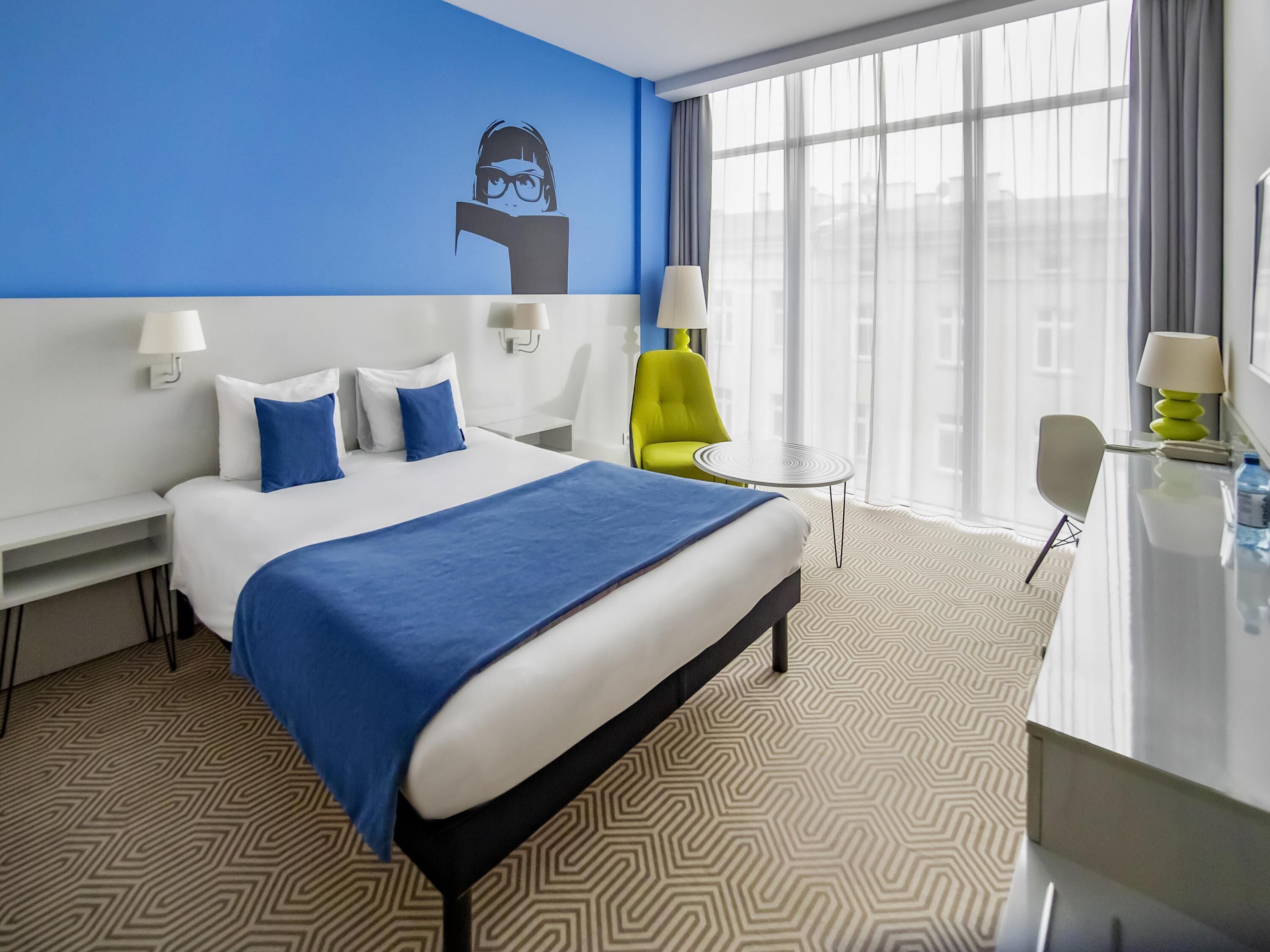 A hotel room with a large bed, a desk, a chair, a small table, and a window with curtains.
