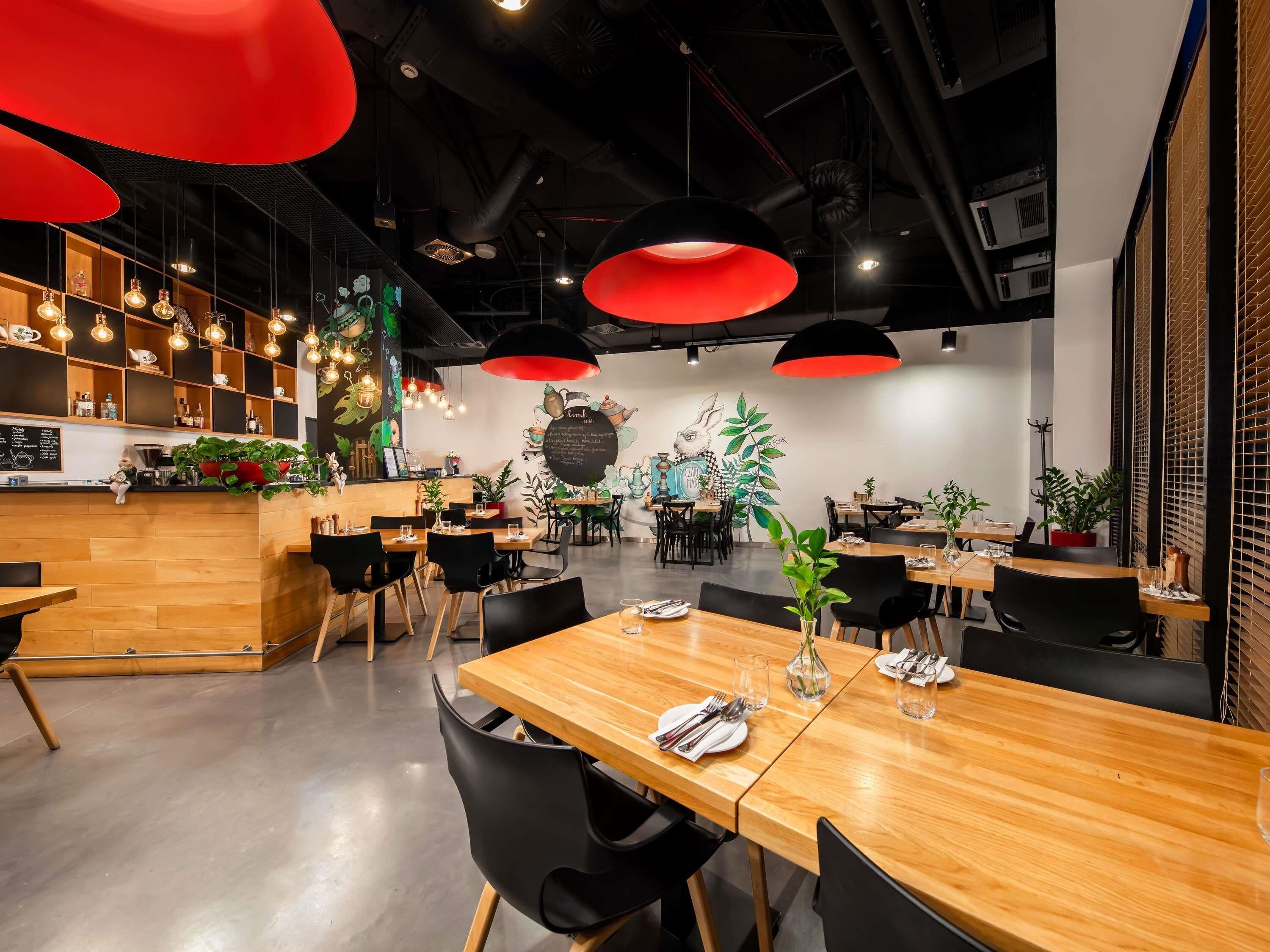 A modern restaurant interior with wooden tables, black chairs, red pendant lighting, and a mural on the wall.
