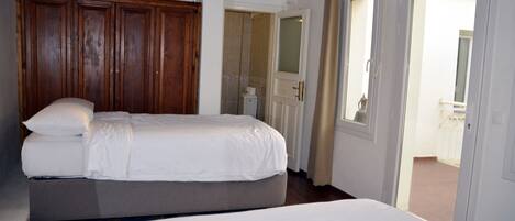A hotel room with two beds, a wooden wardrobe, a bathroom visible through an open door, and a view of the outside balcony.