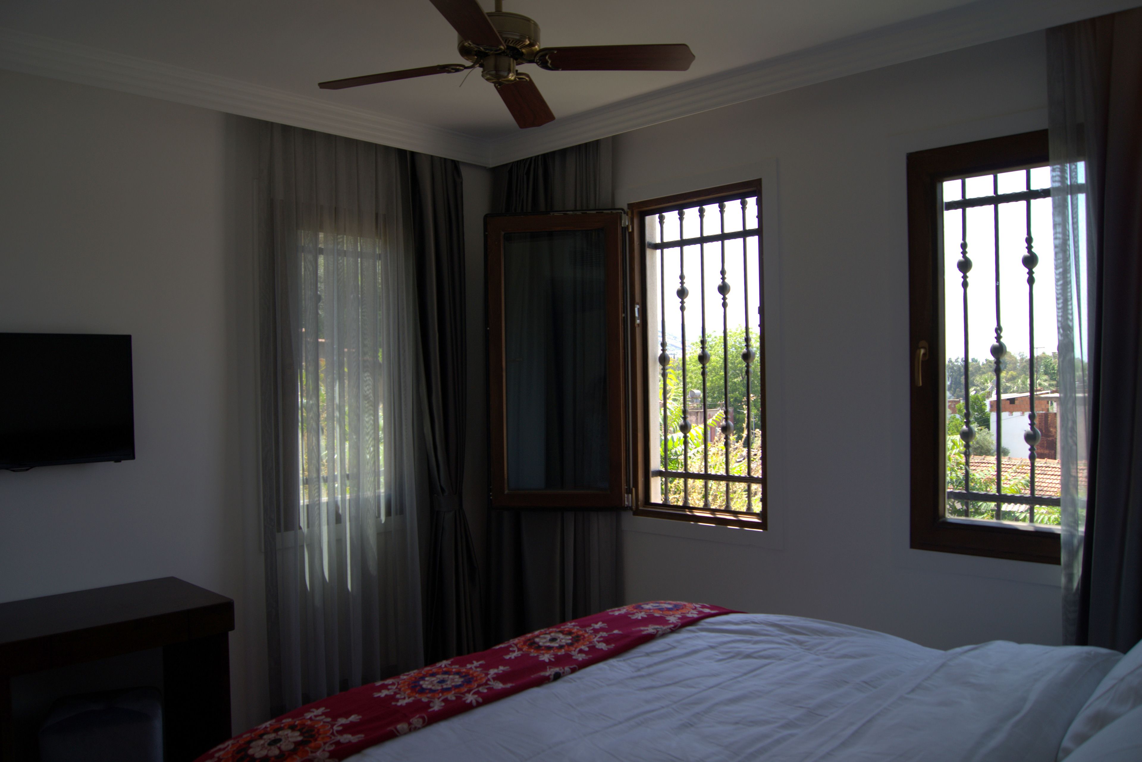 Un dormitorio con cama, ventilador de techo, televisor y ventana con rejas.