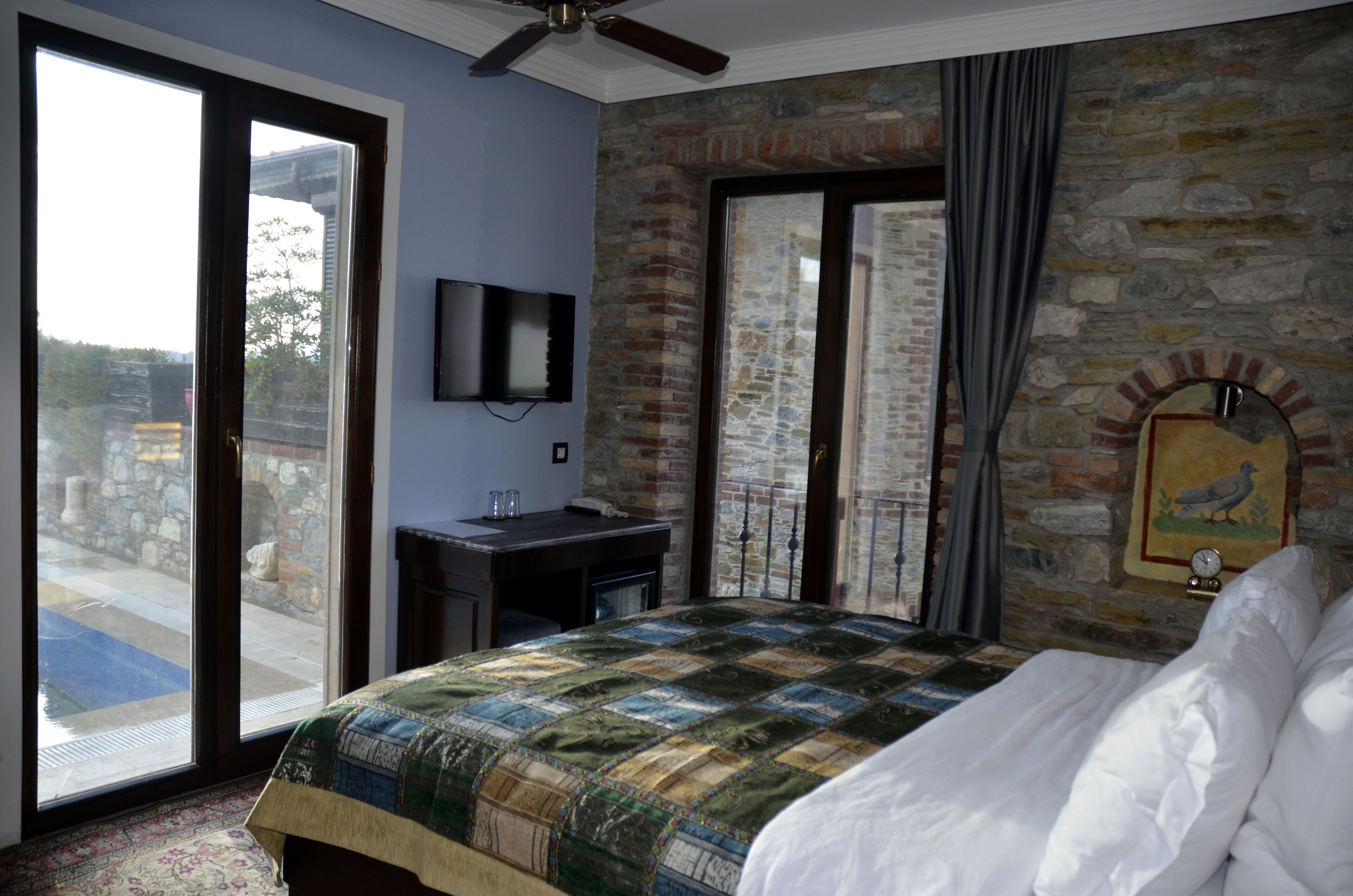 A bedroom with a large bed, a desk, a television, and a stone wall.