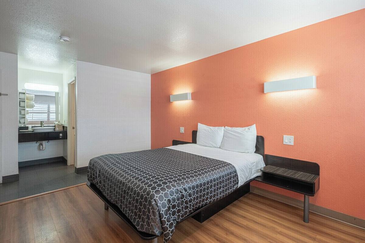Moderni hotellihuone, jossa on sänky, yöpöytä ja kylpyhuone, joka näkyy avoimesta ovesta.