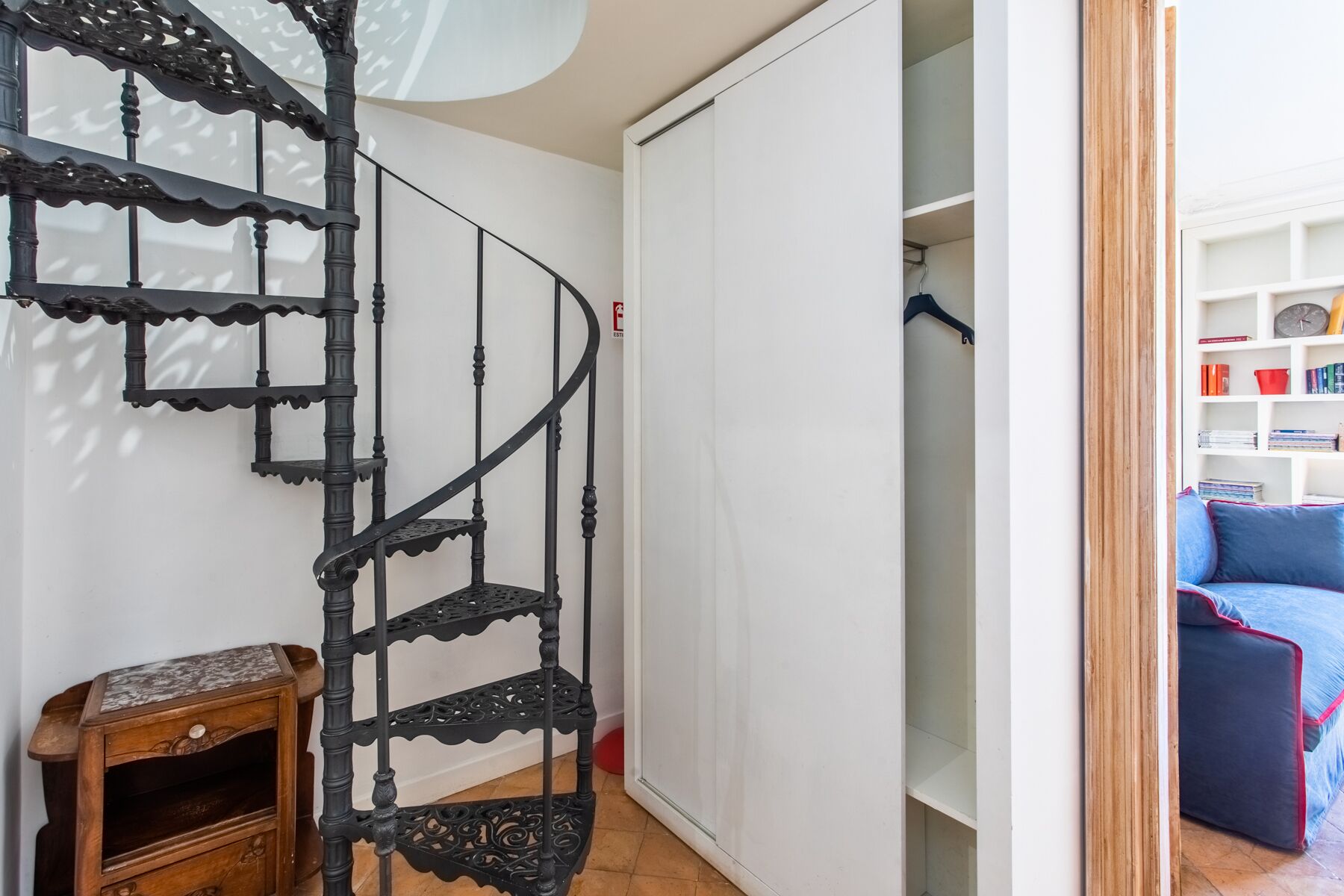 Un escalier en colimaçon orné de rampes noires, une petite table d’appoint en bois et une porte blanche donnant sur une pièce où se trouvent un canapé bleu et une bibliothèque.