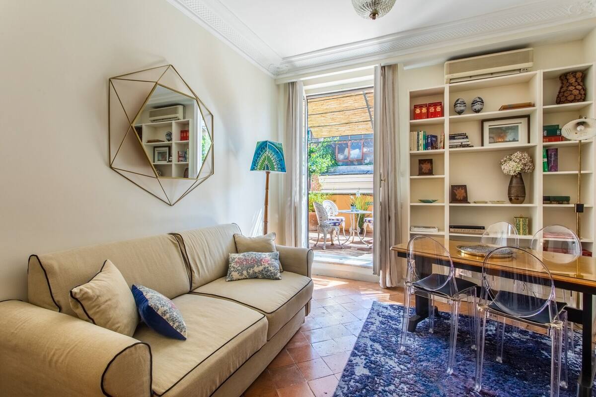 A living room with a beige sofa, a geometric mirror, a wooden dining table with chairs, and a bookshelf.