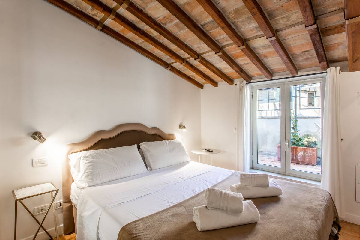 A bedroom with a wooden ceiling, a bed with white linens, a bedside table, and a window with a view of the outside.