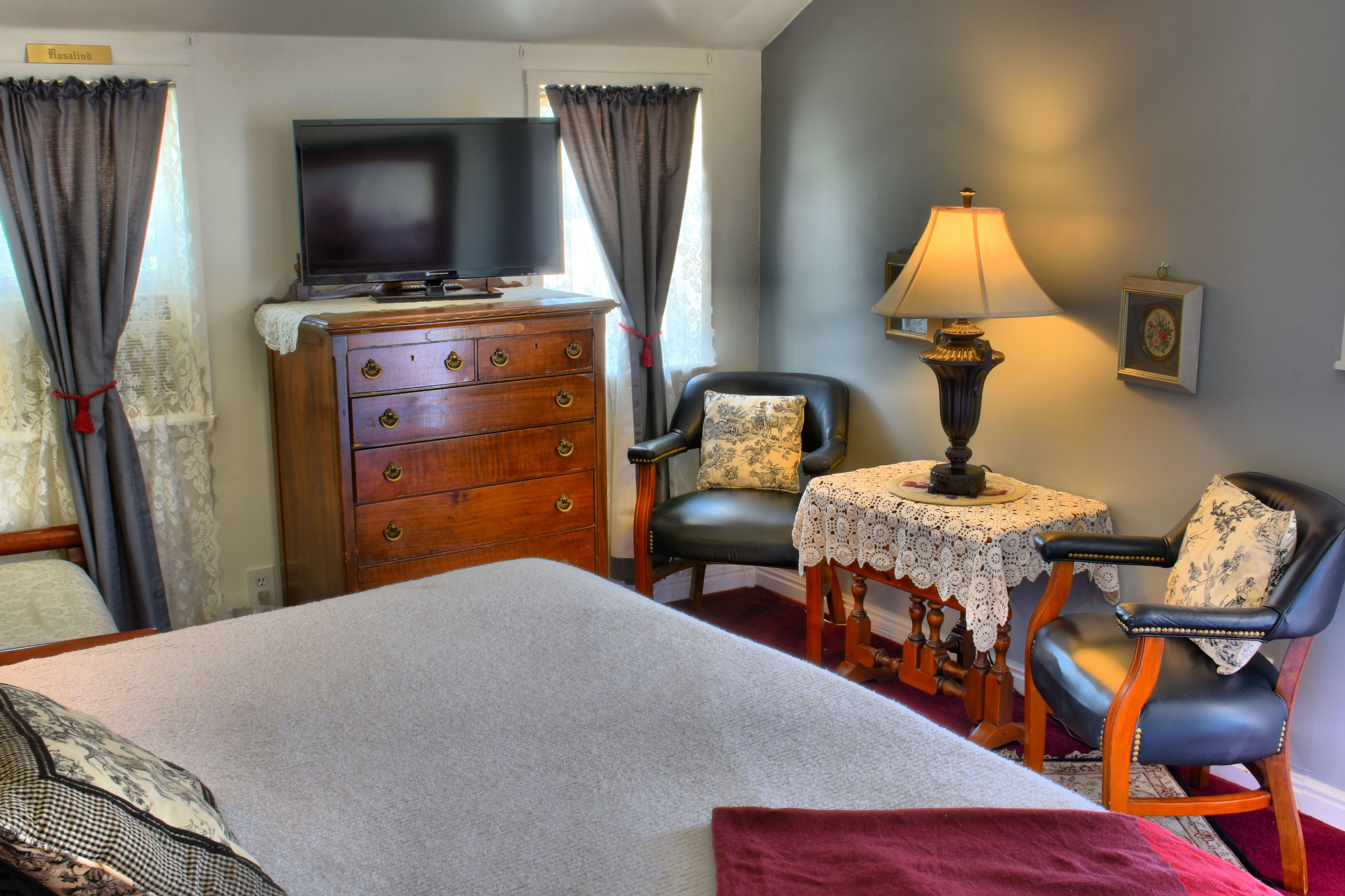 A bedroom with a bed, a dresser, a television, two chairs, a table, and a lamp.